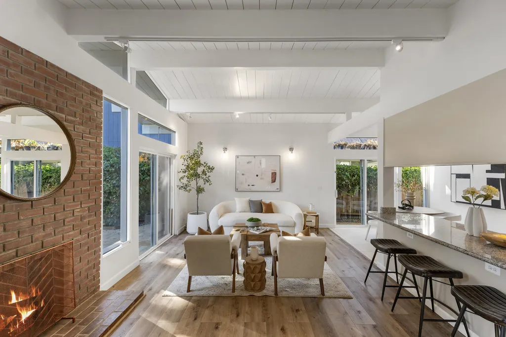 The living room seen past the brick fireplace toward the kitchen counter and dining area, under a beamed ceiling.