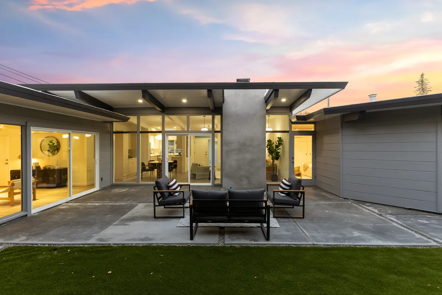 The rear courtyard of an Atherwood Eichler at dusk: glass walls lit from within on three sides beneath a folded roof and a central chimney mass.