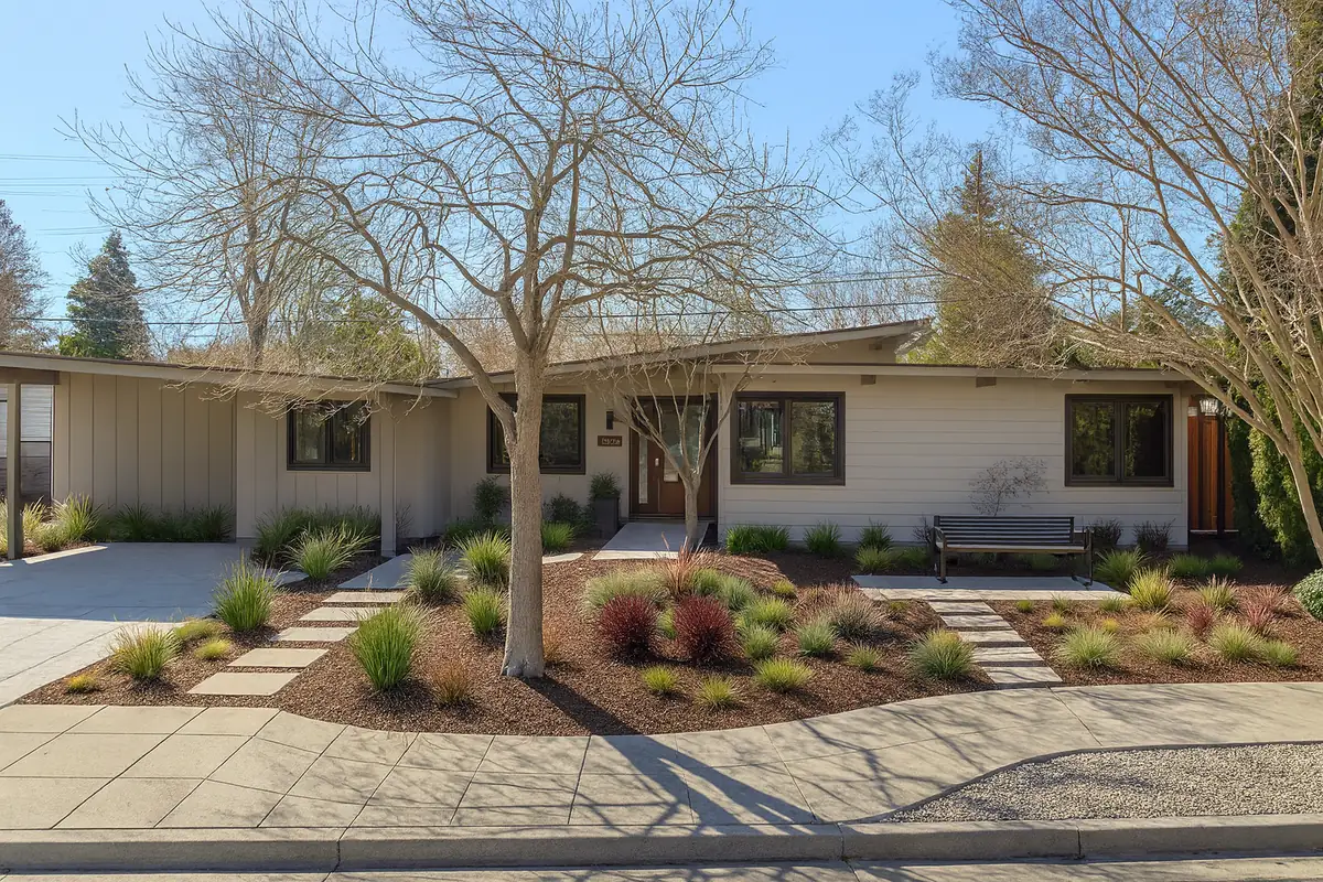 A long single-storey mid-century elevation in white vertical siding, under a bare street tree.