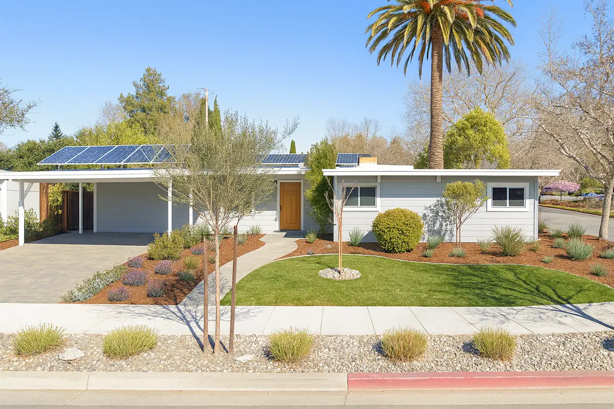 A white single-storey mid-century house with rooftop solar panels above a carport and a low-slope roof.